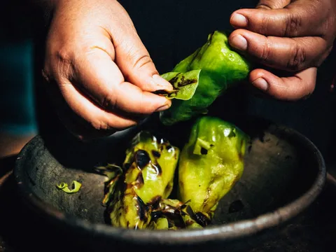 Hands peeling fire-blistered green chiles de agua over a dark clay molcajete at Madre! Oaxacan Restaurant and Mezcaleria