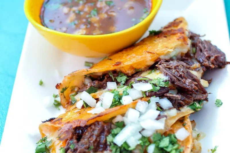 Three crispy quesabirria tacos filled with shredded beef, onion and cilantro on a white plate at Rev Mex, served with a yellow bowl of consommé for dipping