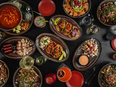 Overhead spread of La Puerta street tacos, loaded fries, pozole, guacamole with chips and margaritas on a dark table