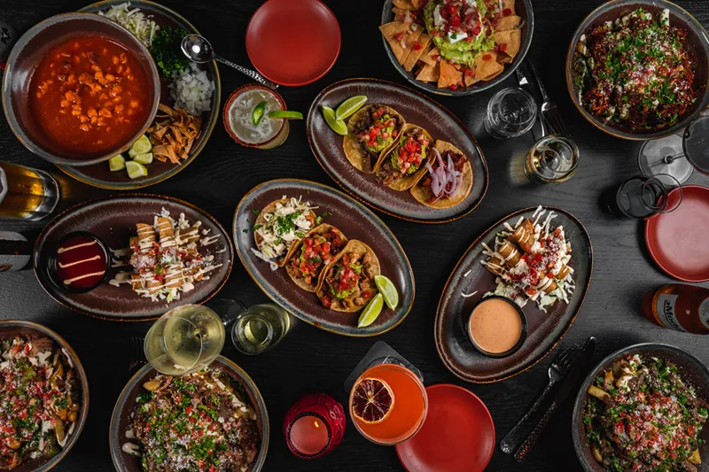 Overhead spread of La Puerta street tacos, loaded fries, pozole, guacamole with chips and margaritas on a dark table