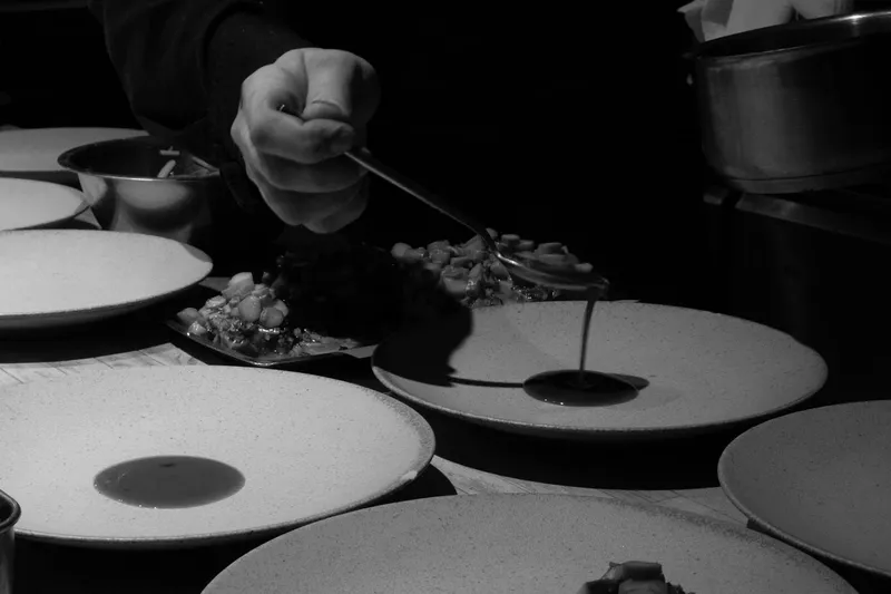 Black-and-white shot of a Corima cook spooning sauce onto speckled ceramic plates of a plated course at the kitchen counter