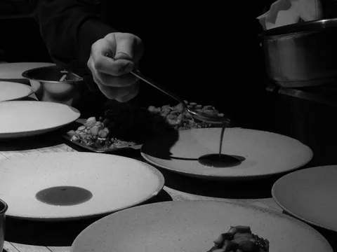 Black-and-white shot of a Corima cook spooning sauce onto speckled ceramic plates of a plated course at the kitchen counter