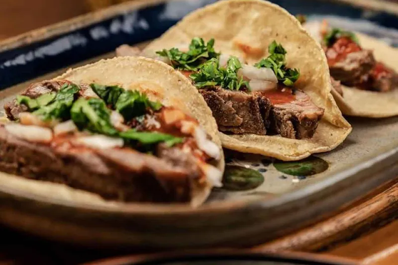 Two bistec tacos on soft corn tortillas topped with cilantro, onion and salsa, served on a blue-rimmed ceramic plate at Mezcal Mexican Cantina