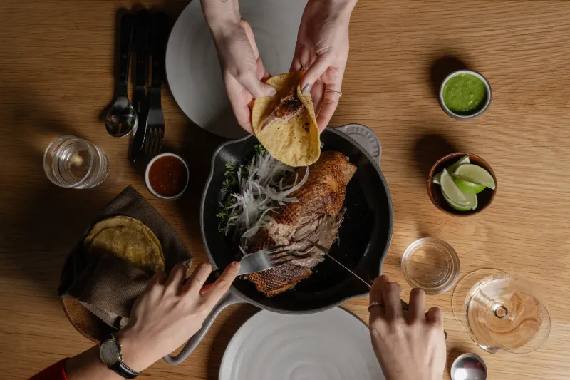 Overhead view of Cosme's Pekin duck carnitas in a cast-iron pan with corn tortillas, salsas and lime for tableside tacos