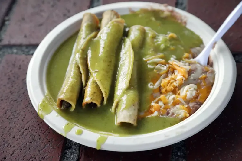 A white plate of Cielito Lindo's rolled beef taquitos smothered in the signature green avocado sauce, with refried beans and shredded cheese, set on an Olvera Street brick surface