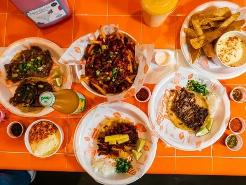 An overhead spread of Taquerias Carnalito dishes on an orange-tiled table — carne asada tacos, al pastor tacos, loaded Desmadre fries, tortilla chips with queso, elote, salsas and a Boing bottle