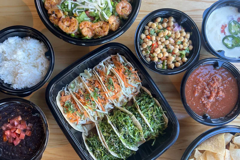 Overhead spread of Taqueria Tsunami fusion tacos, a grilled shrimp bowl, rice, black beans, chickpea salad, queso, salsa and tortilla chips in black takeout containers
