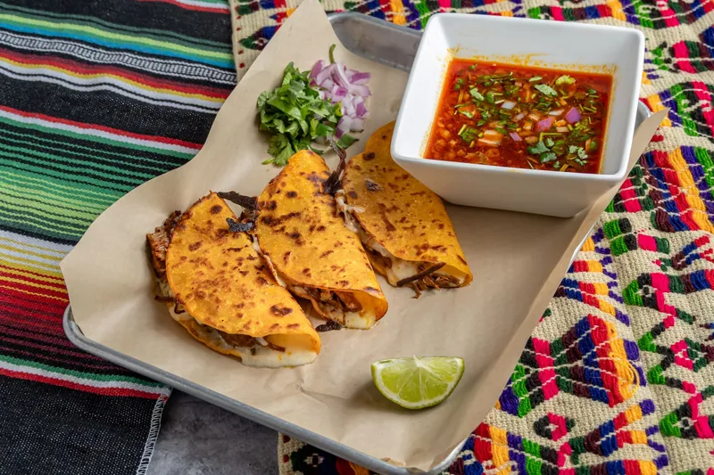An order of Taqueria Number 1 quesabirria tacos — crispy cheese-filled tacos with a cup of red beef consomé, chopped onion and cilantro, on a paper-lined tray