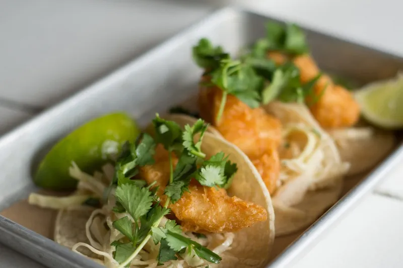 Crispy battered shrimp tacos topped with cilantro, cabbage slaw and lime wedges on a metal tray at Bartaco