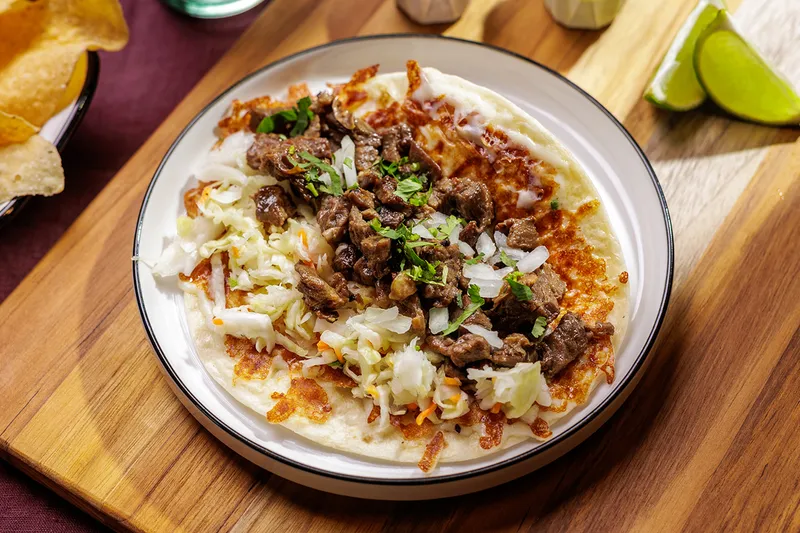 A Salsa Limón taco 'con todo' — grilled asada with crispy melted cheese, cabbage, onion and cilantro on a flour tortilla, served with lime wedges and tortilla chips on a wooden board