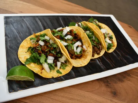A plate of four Basilios Tacos street tacos — soft corn tortillas topped with grilled meat, chopped onion and cilantro — with a lime wedge, on a dark rectangular plate over a wood table