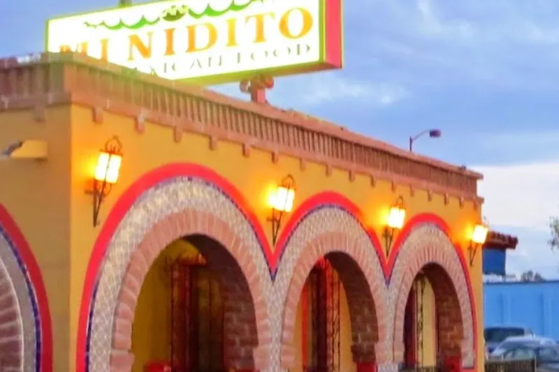 The illuminated 'Mi Nidito Mexican Food' rooftop sign above the colorful yellow, red and tiled arched storefront of Mi Nidito Restaurant in South Tucson at dusk