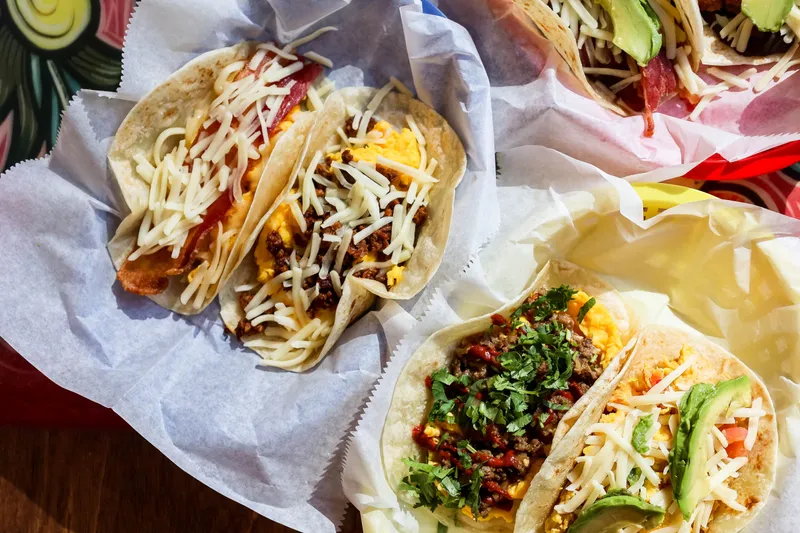 A paper-lined basket of Tacodeli breakfast tacos on corn and flour tortillas — bacon and egg, chorizo and picadillo with shredded cheese, cilantro and avocado